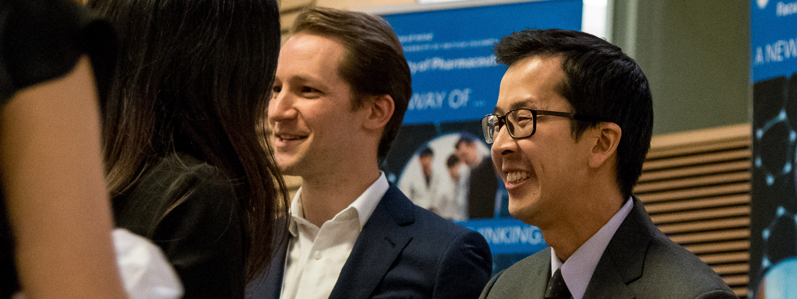 A group of people mingling at an event, with two men in focus and two banners in the background.