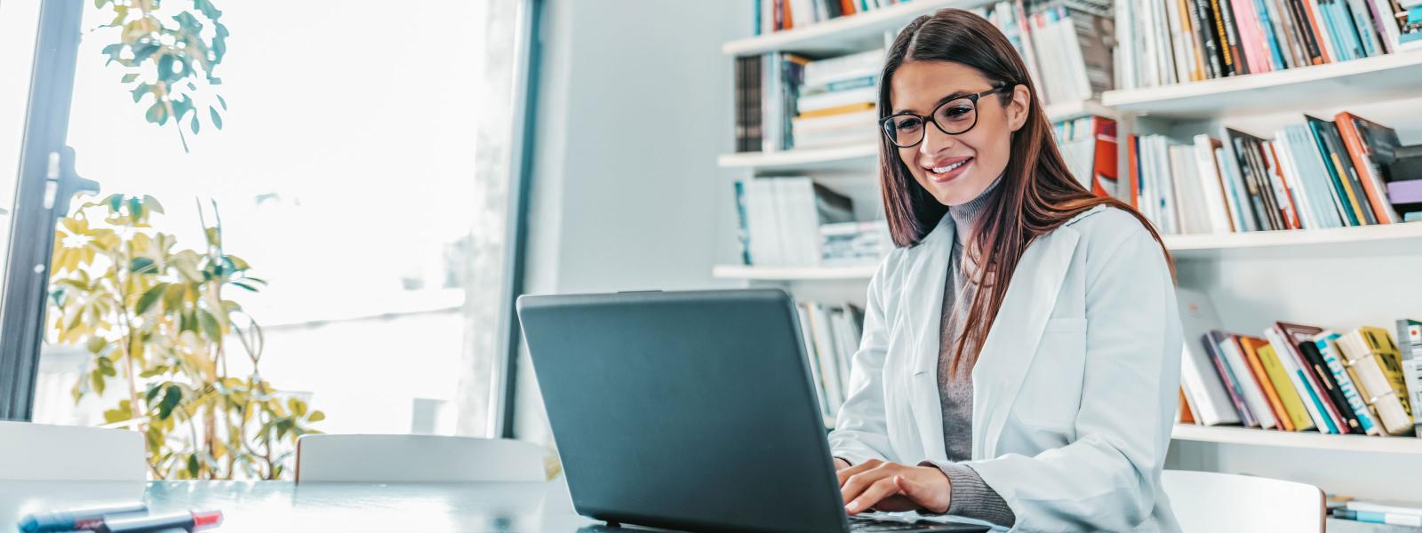 A health care practitioner smiling as she works at a laptop in a well-lit room.
