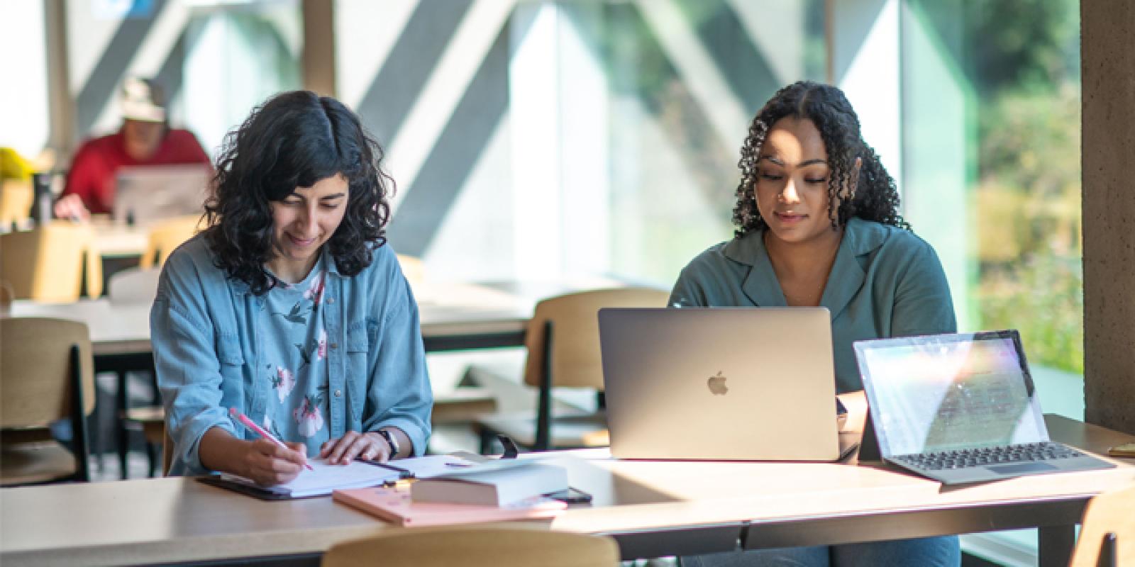 Two students working at a shared desk, one with a notebook and one with a laptop.