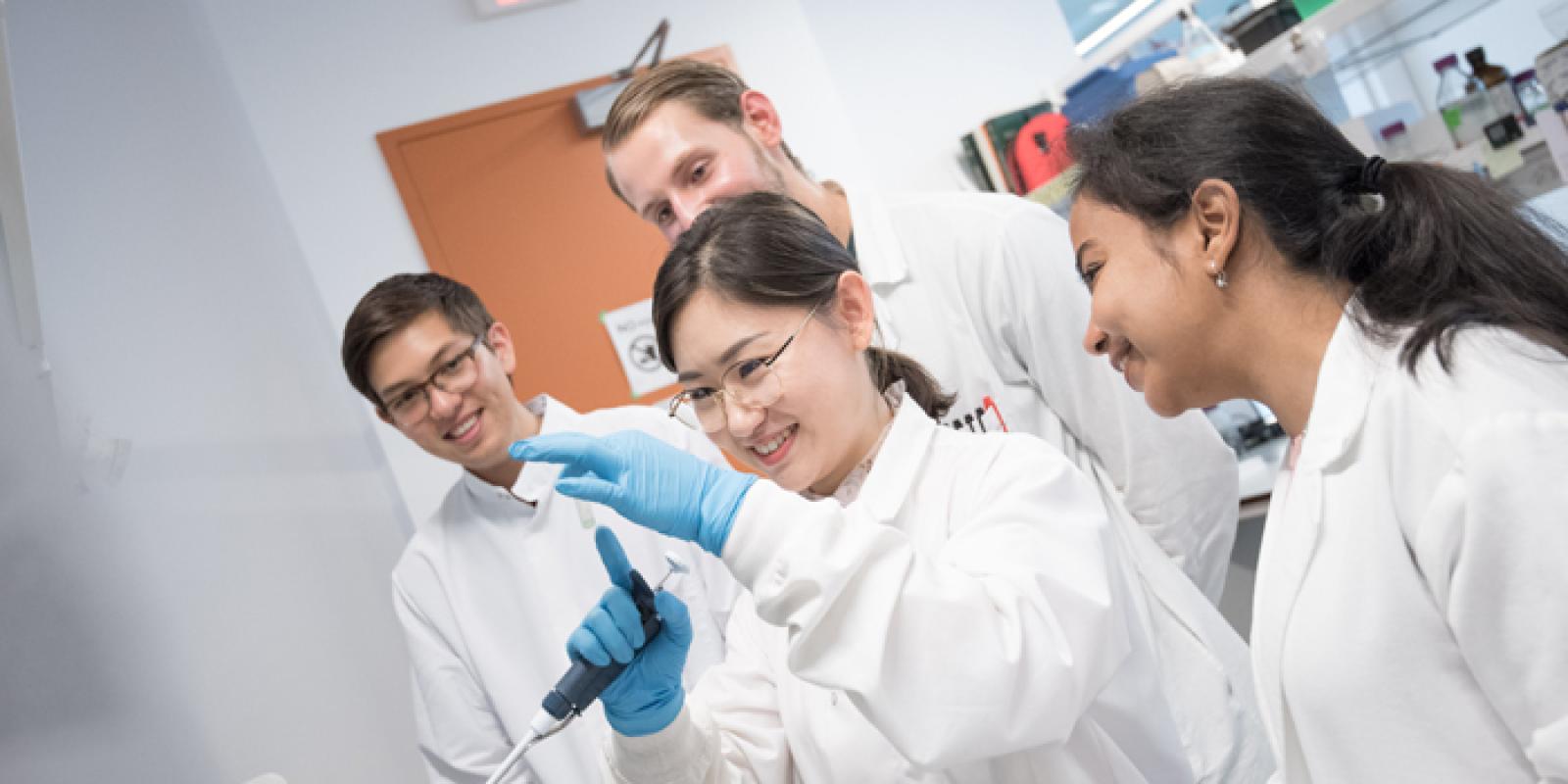 Four students wearing lab coats in a lab; the student in the middle is holding some lab equipment and smiling while the others look on.