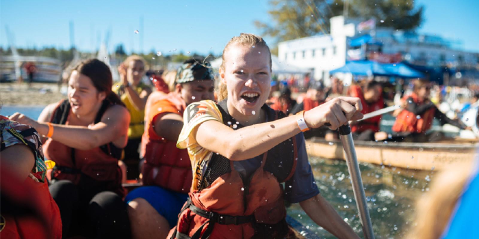 A group of students smiling while they practice rowing a longboat on a sunny day. 
