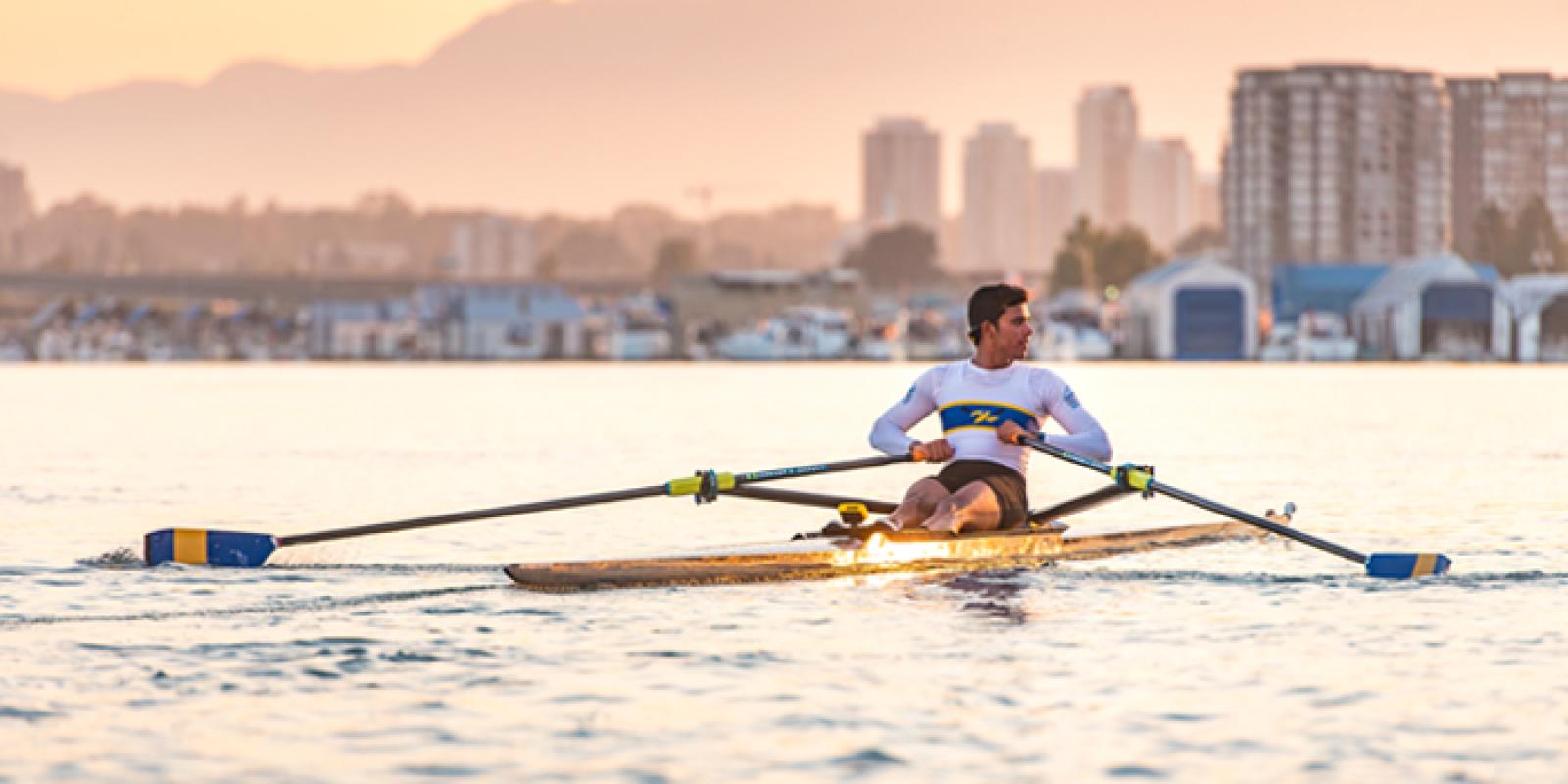A student rowing a boat at sunset, with a city skyline in the distance.