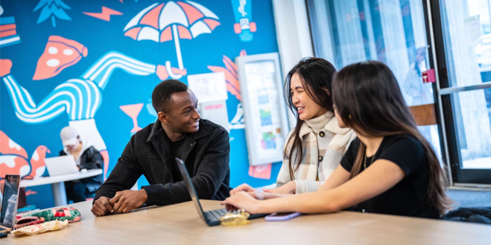 Three students conversing and smiling at a table.