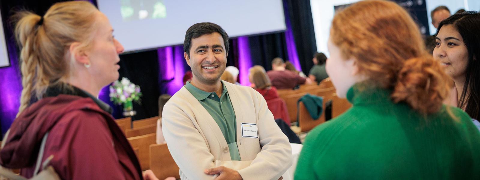 Four people conversing at an alumni event, with more people mingling in the background.