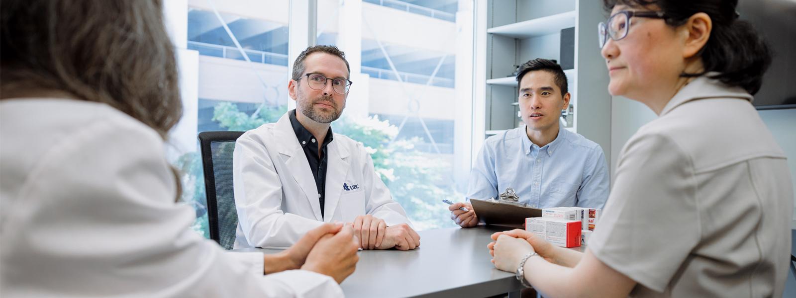 Four people sitting and conversing at a desk, two of whom are wearing lab coats.
