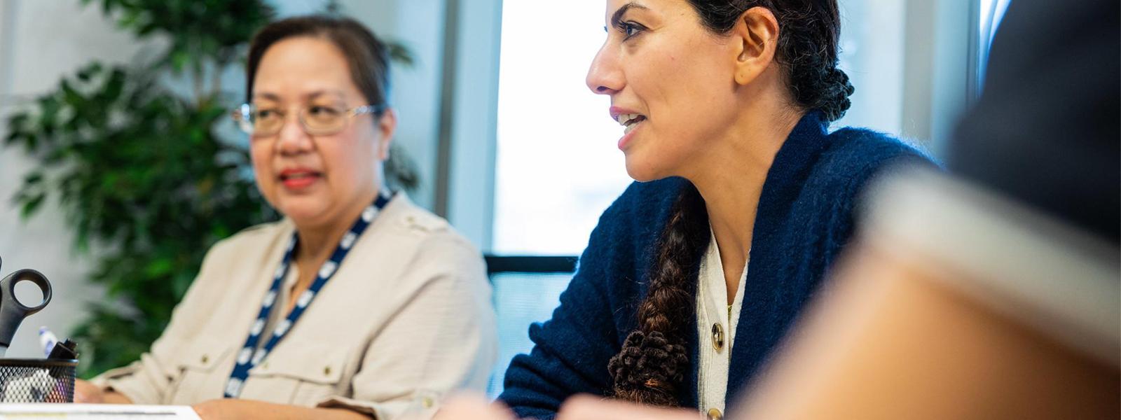 Two women sitting and speaking to others offscreen in a meeting room.