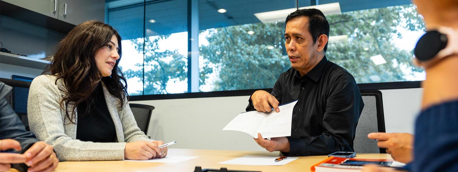 A man gesturing at a brochure while a woman, and others offscreen, look on in a meeting room.