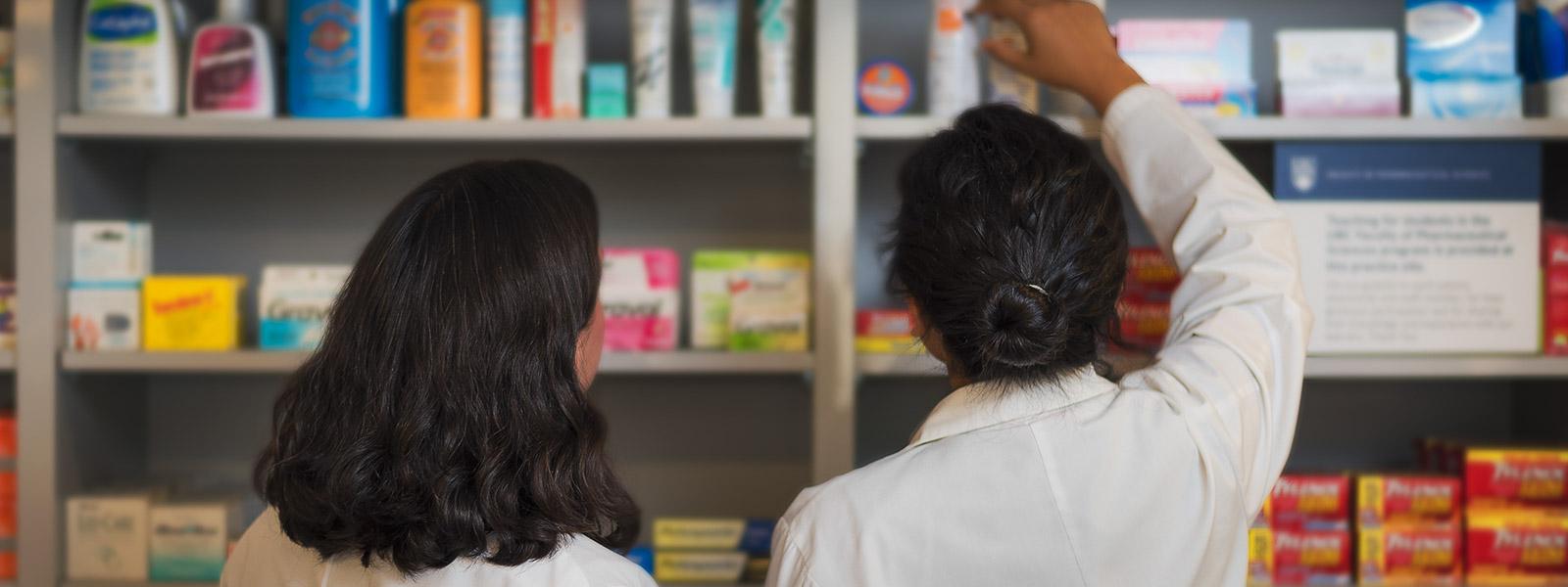 Two pharmacists facing away from the camera, looking at a shelf of medications.