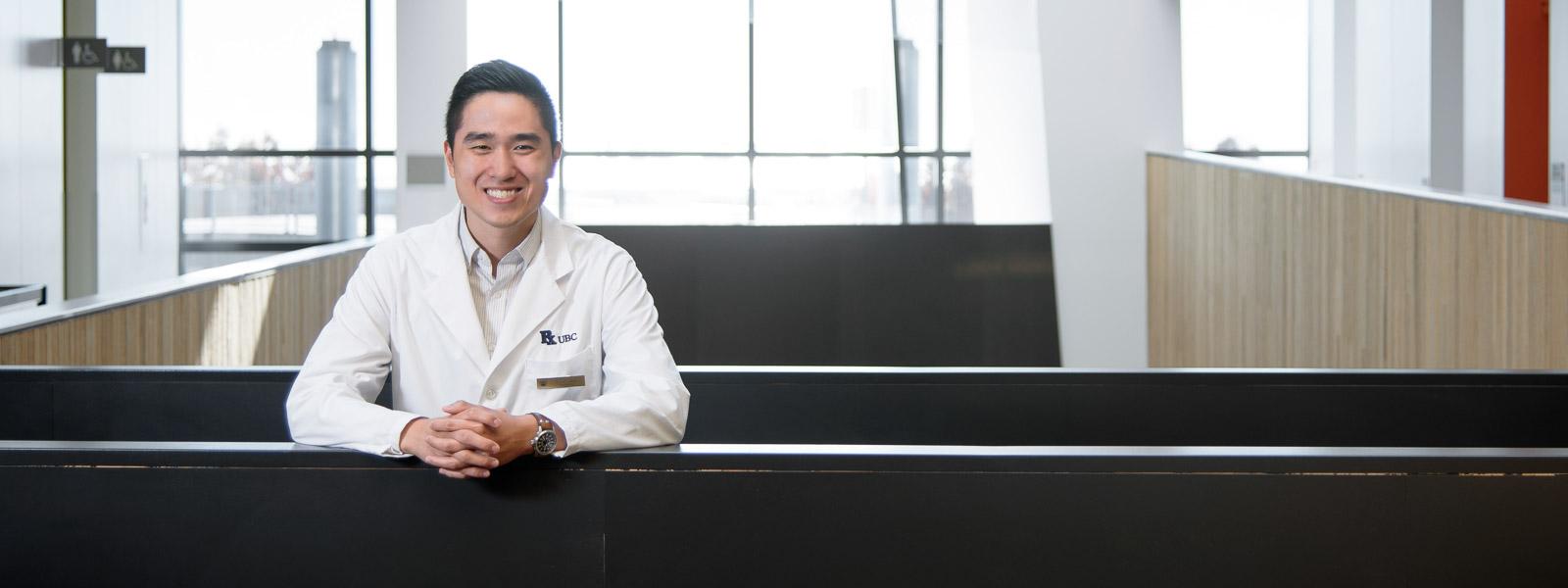 A man in a lab coat, leaning on a railing inside the Pharmaceutical Sciences Building, smiling at the camera.
