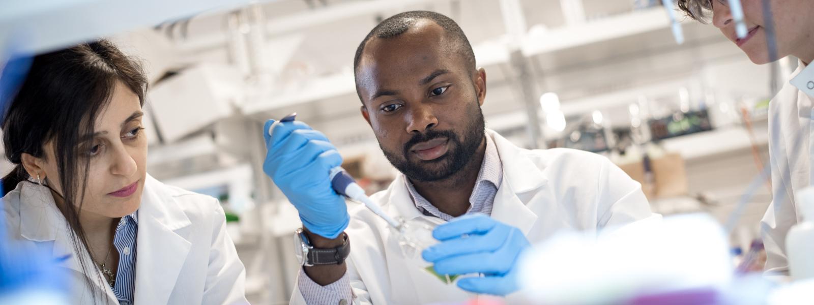 Three people in a lab; the man in the middle is concentrating on the equipment he is holding.